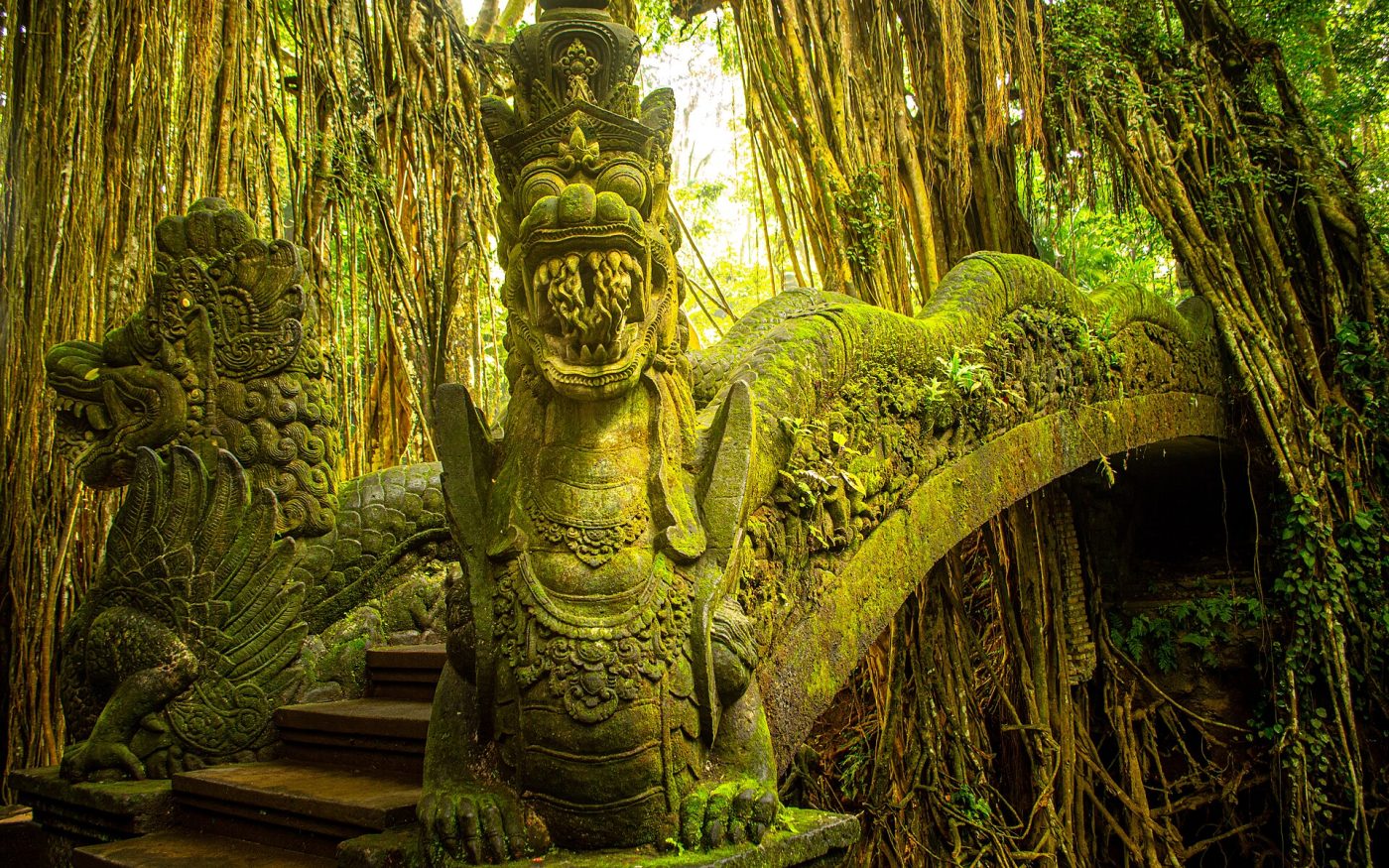 Moss-covered dragon bridge and stone statues under banyan roots in the Sacred Monkey Forest, Ubud