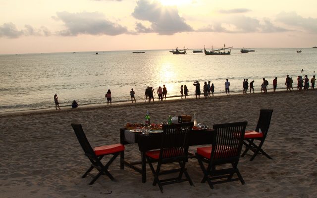 Table for dinner set on the sand at Jimbaran Beach with fishing boats and the sunset over the bay