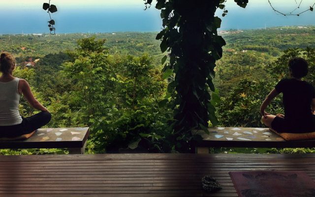 Two people meditating on a wooden yoga deck overlooking jungle and the sea in Bali