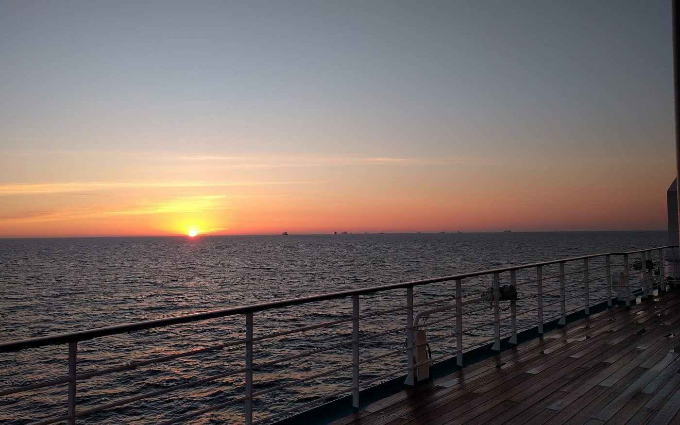 Orange sunset over a calm sea seen from the wooden deck and railing of a cruise boat