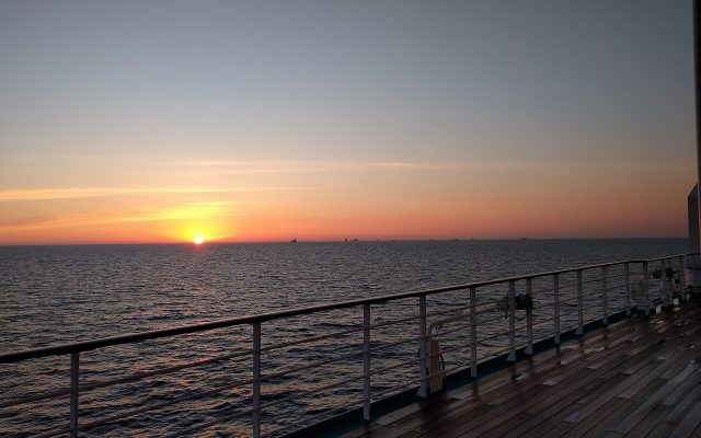 Orange sunset over a calm sea seen from the wooden deck and railing of a cruise boat