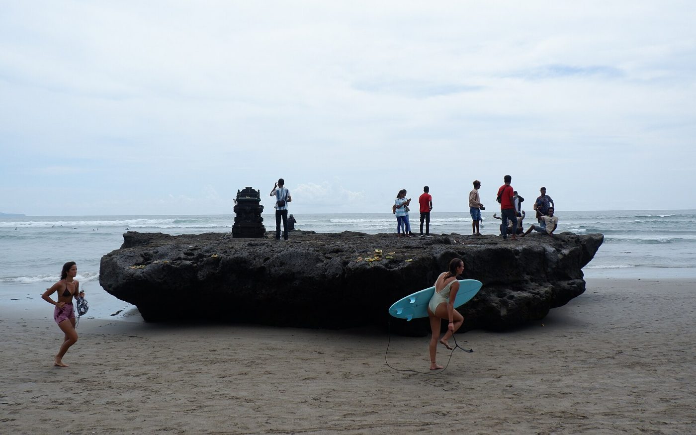 Surfer carrying a board across the sand past the shrine rock at Canggu Beach, Bali