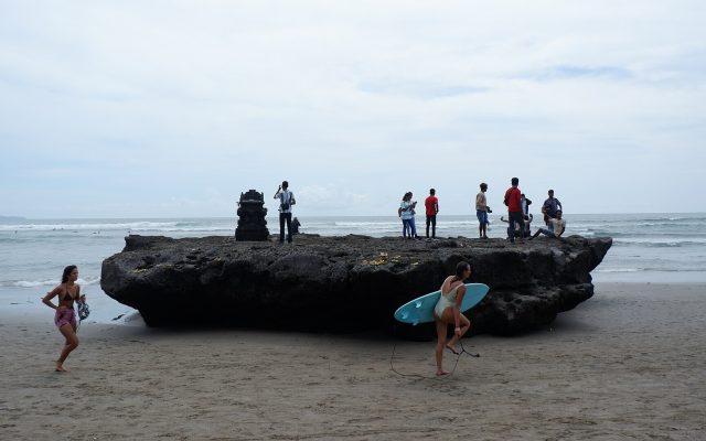 Surfer carrying a board across the sand past the shrine rock at Canggu Beach, Bali