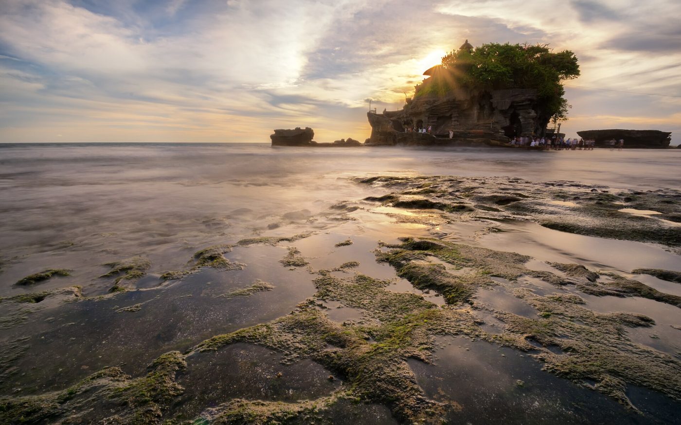 Sun rays behind Tanah Lot temple with the rocky shore in the foreground at sunset
